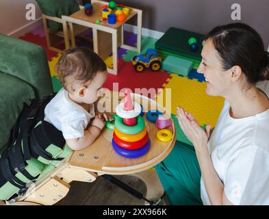 Il bambino con ipotonia corporea sta lavorando su attrezzature per la riabilitazione in piedi con fisioterapista utilizzando anelli colorati impilabili in un bambino-fr Foto Stock