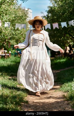 Una giovane donna in abito bianco e cappello di paglia cammina lungo un sentiero in un giardino estivo. Foto Stock