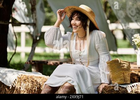 Una giovane donna con un abito bianco e un cappello di paglia sorride brillantemente mentre si siede in un giardino estivo, godendosi il sole. Foto Stock