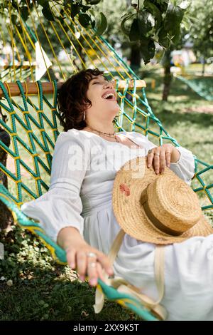 Una donna con un abito bianco e un cappello di paglia ride gioiosamente mentre si rilassa su un'amaca in una soleggiata giornata estiva. Foto Stock