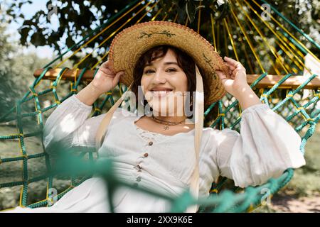 Una donna bruna si rilassa in un'amaca, indossando un abito bianco e un cappello di paglia, godendosi il calore di una giornata estiva. Foto Stock