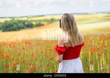 Una giovane bionda scatta una foto con un telefono in un campo di papaveri rossi Foto Stock