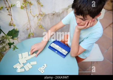 Un ragazzo si siede con cura mentre gioca a domino su un tavolo blu all'esterno. Ama la calma e la concentrazione del gioco in un ambiente naturale. Foto Stock