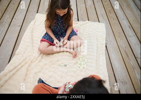 Due bambini seduti su una coperta calda, impegnati in un divertente gioco di domino su una terrazza soleggiata all'aperto, godendosi del tempo libero. Foto Stock