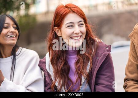 Primo piano di una giovane donna gioiosa con i capelli rossi, sorridente in generale mentre passava del tempo con il suo gruppo eterogeneo di amici. Ritratto autentico di h Foto Stock