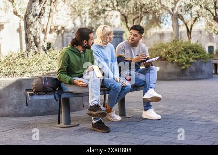 Gruppo di giovani studenti diversi che collaborano nel campus universitario. Tre amici di diverse etnie seduti sulla panchina, a discutere e a studiare Foto Stock