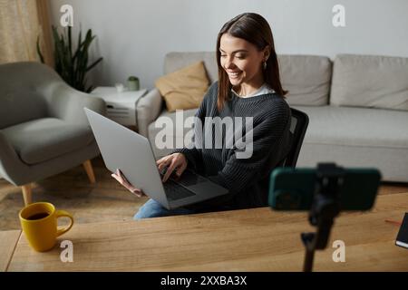 Una giovane donna con un maglione grigio lavora sul suo notebook a una scrivania mentre filma un video. Foto Stock