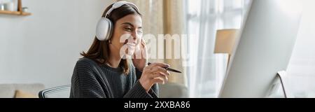 Una giovane donna lavora in remoto da casa, indossando le cuffie e un maglione grigio mentre crea contenuti sul computer. Foto Stock