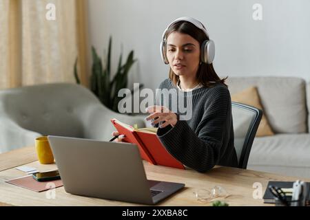 Una donna che indossa le cuffie lavora al notebook, tenendo aperto un notebook rosso mentre guarda lo schermo. Foto Stock