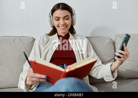 Una giovane donna con un abbigliamento casual si rilassa sul divano, ascoltando musica con le cuffie wireless e leggendo un libro rosso. Foto Stock