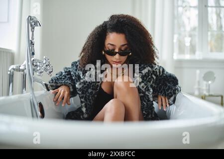 Un'elegante donna afroamericana con un cappotto con stampa leopardata si posa in una vasca da bagno, con le gambe incrociate e gli occhiali da sole appoggiati sul naso. Foto Stock