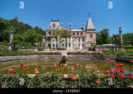 Una grande casa con una fontana di fronte. La fontana è circondata da fiori rossi. La casa è circondata da un giardino con molti alberi e. Foto Stock