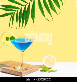 Cocktail estivo blu con menta e limone su sfondo giallo brillante con foglie di palma. Concetto creativo alla moda. Cocktail laguna blu con liquore curacao Foto Stock