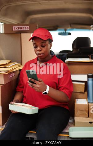 Donna afroamericana seduta tra pacchi e scatole mentre utilizza il telefono cellulare per aggiornamenti sullo stato della spedizione Foto Stock