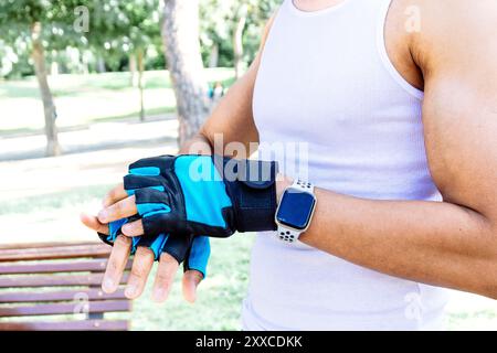 Un uomo con canotta bianca e pantaloncini neri indossa un cinturino da polso blu. E' in piedi in un parco ed e' concentrato su qualcosa Foto Stock