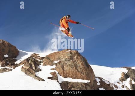 Uno sciatore professionista fa un salto da un'alta scogliera contro il cielo blu lasciando una scia di neve in montagna. Foto dalle piste o Foto Stock