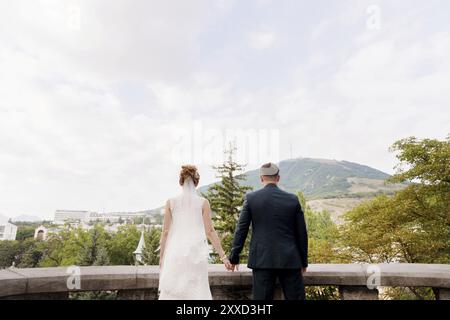 Una coppia amorevole di sposi novelli sta con la schiena che si tiene per mano e si abbraccia sullo sfondo del paesaggio urbano della città del resort a nord Foto Stock