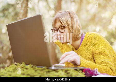 Immagine tonificata di un ritratto di una ragazza freelance con un maglione giallo e occhiali che guardano con attenzione lo schermo del notebook nella natura in un fronte di conifere Foto Stock