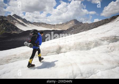 Un alpinista con uno zaino cammina in ramponi camminando lungo un ghiacciaio polveroso con marciapiedi tra le fessure della montagna Foto Stock