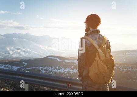 Uomo hipster turistico barbuto con occhiali da sole e zaino, stenditi su un dosso lungo la strada e guarda il tramonto sullo sfondo di una cima innevata Foto Stock
