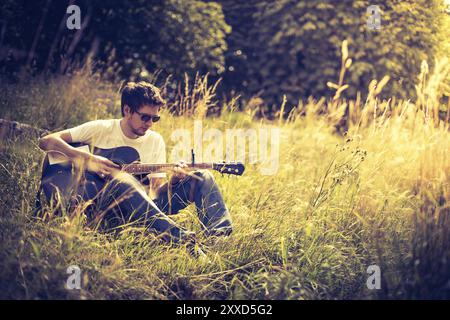 Il giovane è seduto su un fiume e gioca il suo chitarra occidentale Foto Stock