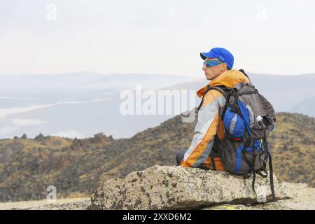 Un turista solitario con zaino e bastoncini per muoversi in montagna. In occhiali da sole e con uno zaino poggiare la sidiya su una pietra. Vista dal bac Foto Stock