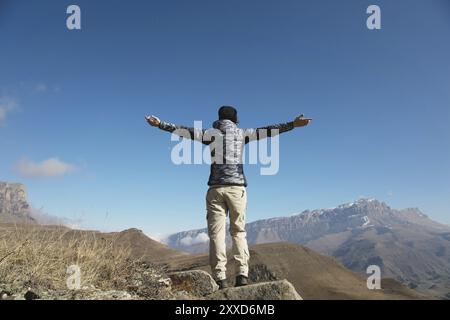 Un applauso di una giovane donna in spalla sulla cima di un'ampia cima di montagna. Libertà e vittoria sullo sfondo di montagne e blu Foto Stock