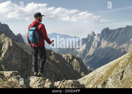 Un uomo barbuto con occhiali da sole e un berretto con uno zaino si staglia sulla cima di una roccia e guarda verso una valle rocciosa in alto tra le montagne. Il concetto di tour Foto Stock