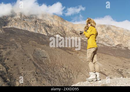 Una fotografa con occhiali da sole e un cappello di pelliccia grande e un maglione in maglia gialla si staglia sullo sfondo di alte rocce nella gola con una cameriera Foto Stock