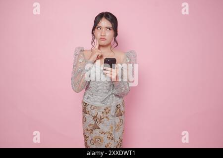 Giovane donna indonesiana premurosa che indossa un moderno kebaya tenendo in mano lo smartphone mentre pensa a fondo rosa isolato. Foto Stock