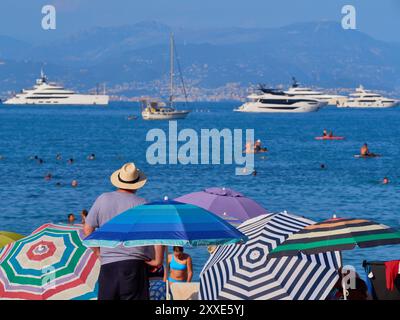Scenario di spiaggia con un turista in cappello di paglia che guarda yacht e superyacht ancorati nella baia di Antibes, Costa Azzurra, Francia. Foto Stock