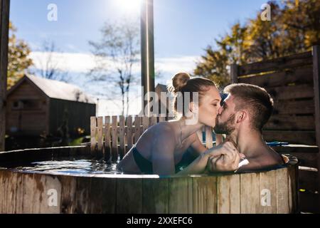 Dolce bacio di una giovane coppia che si diverte e si rilassa in un'area spa privata, una vasca idromassaggio all'aperto in un resort glamping. Foto Stock