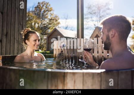 Giovane coppia attraente che gode di una vasca idromassaggio privata in legno durante una vacanza glamping. Concetto di fuga romantica per il weekend. Foto Stock