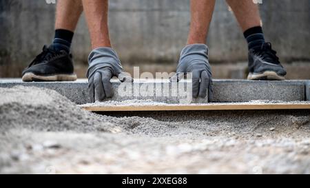 Visuale ad angolo basso di un operatore con guanti protettivi che posa piastrelle di cemento all'esterno per realizzare una nuova pavimentazione. Foto Stock