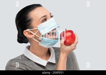 Una giovane donna con maschera mangia una mela in ufficio. Assistenza sanitaria. Foto Stock