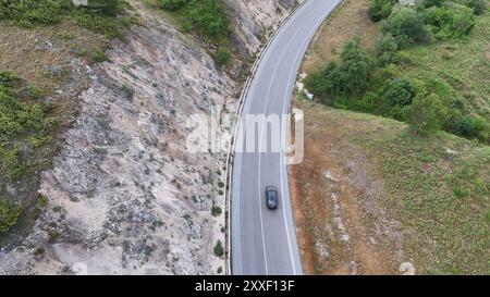 Un'auto elegante scivola lungo una strada curva che abbraccia un'aspra scogliera, circondata da vegetazione lussureggiante e dall'atmosfera serena di una giornata coperta. Foto Stock