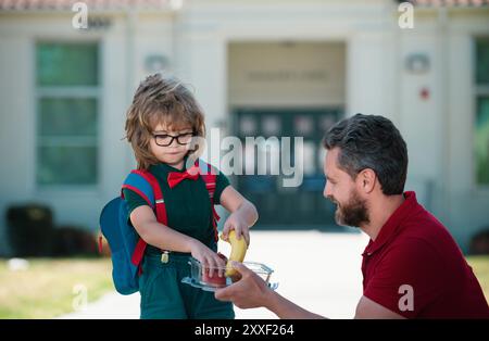 Il Padre sostiene e motiva il figlio. Capretto che va alla scuola primaria. Piccolo scolaro che mangia gustoso pranzo all'aperto. Giornata degli insegnanti. Foto Stock