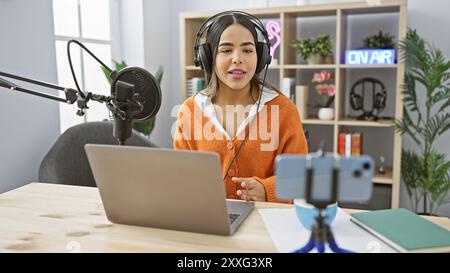 La giovane donna ispanica ospita podcast in uno studio radio interno con microfono e laptop Foto Stock