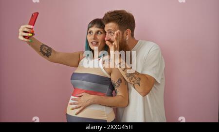 Donna incinta e uomo che fanno un selfie insieme a uno sfondo rosa che mostra il loro amore e i loro tatuaggi Foto Stock