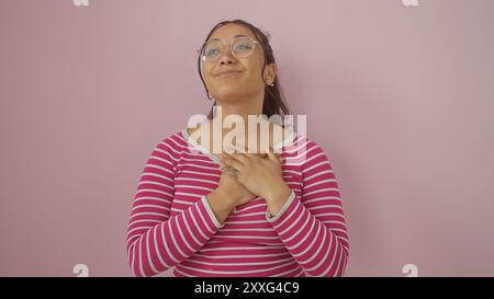 Giovane donna ispanica sorridente e che mette le mani sul cuore, indossa occhiali e una camicia a righe su sfondo rosa. Foto Stock