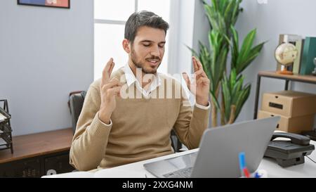 Uomo ispanico a dita incrociate con maglione e camicia che lavora al suo laptop in un ufficio moderno. Foto Stock