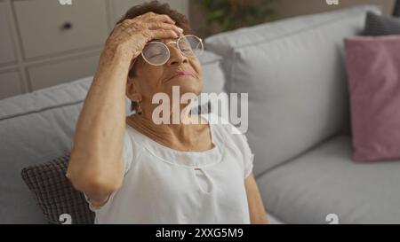 Anziana donna ispanica con capelli corti e occhiali che toccano la fronte mentre è seduta nel salotto di casa sua. Foto Stock