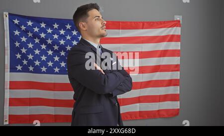 Un giovane ispanico sicuro di sé con una tuta si erge con le braccia incrociate in un ufficio con una bandiera americana sullo sfondo. Foto Stock