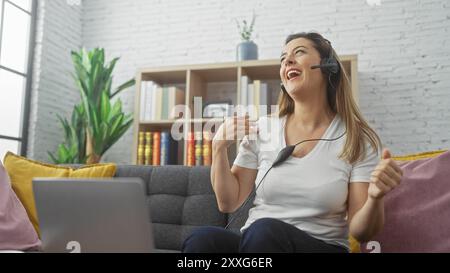 La donna sorridente con la cuffia utilizza il computer portatile in un salotto accogliente, esprimendo positività e comfort a casa. Foto Stock