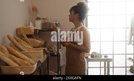 Donna che lavora in una panetteria che ispeziona dolci con un appunti, raffigurante un giovane, bello, adulto, ispanico, bruna con capelli ricci in una s Foto Stock