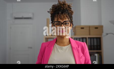 Giovane donna in ufficio con capelli ricci che indossa blazer rosa e occhiali che sorride con fiducia Foto Stock