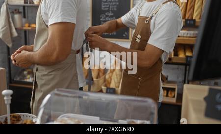 Fornai maschili che lavorano insieme in una panetteria interna, regolando i grembiuli e preparandosi per il servizio, circondati da pane e dolci freschi Foto Stock