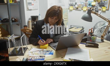 Detective donna matura che scrive appunti in un ufficio del dipartimento di polizia circondato da indizi. Foto Stock