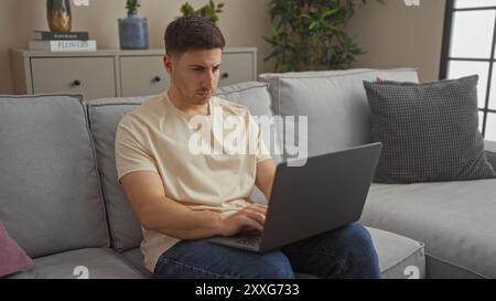 Giovane ispanico che lavora su un computer portatile in un salotto accogliente, seduto su un comodo divano, con piante e decorazioni sullo sfondo, creando un Foto Stock