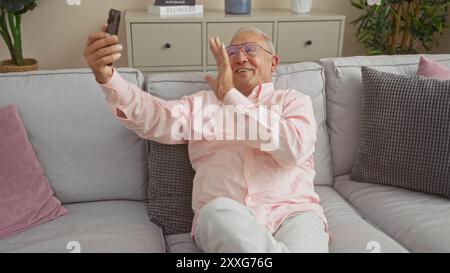 Un uomo anziano con i capelli grigi che indossa gli occhiali e una camicia rosa scatta un selfie mentre sorride e sventola in un accogliente soggiorno. Foto Stock